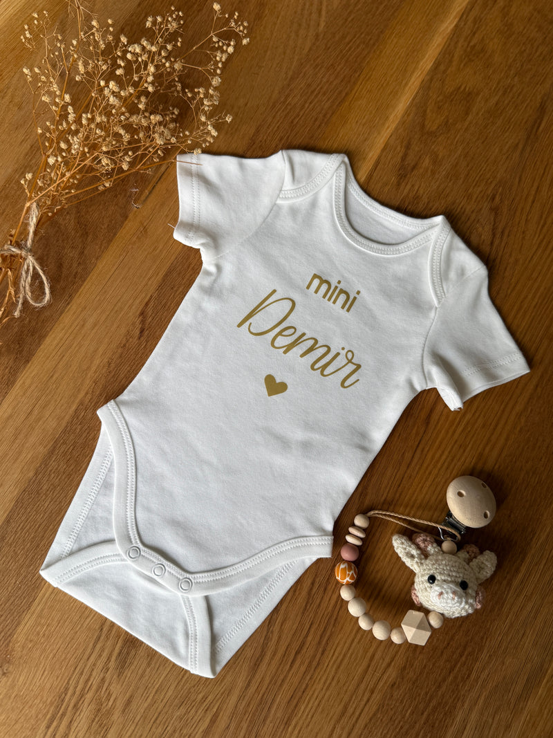 Personalisierbarer Babybody aus Bio-Baumwolle mit goldfarbener Schrift und Herzmotiv.