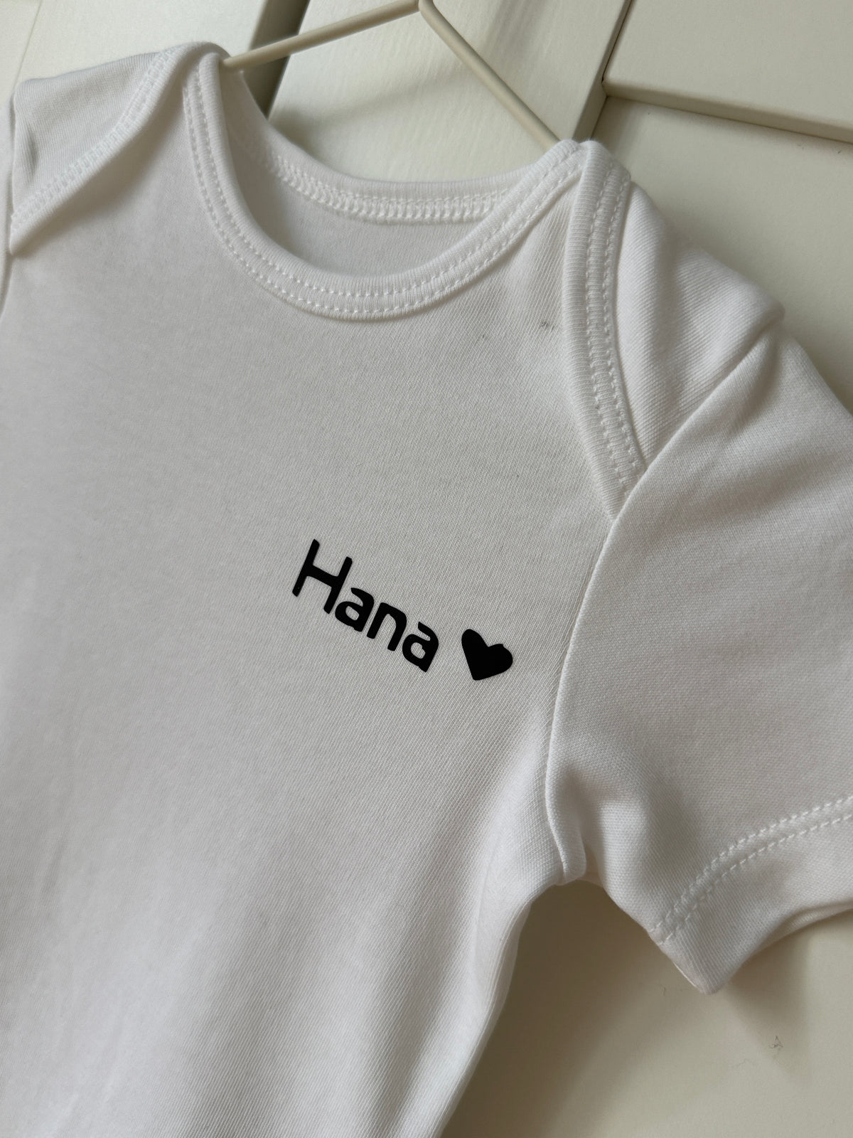 Weisser Baby-Body aus Bio-Baumwolle personalisiert mit dem Namen 'Hana' und einem Herz-Symbol.